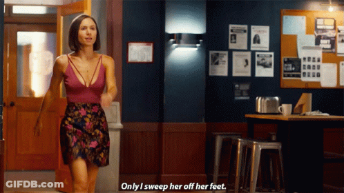 A Woman In A Floral Skirt Says " Only I Sweep Her Off Her Feet " In Front Of A Bulletin Board GIF