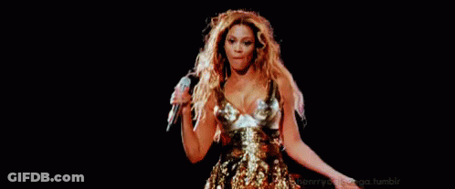 A Woman In A Gold Dress Is Singing Into A Microphone On A Stage In Front Of A Black Background . GIF