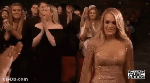 A Woman In A Gold Dress Is Standing In Front Of A Crowd Of People Clapping . GIF