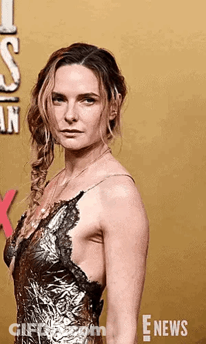 A Woman In A Gold Dress Is Standing On A Red Carpet And Looking At The Camera . GIF