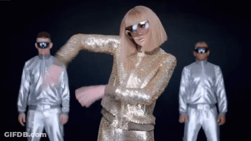 A Woman In A Gold Sequined Outfit Is Dancing In Front Of Two Men In Silver Suits GIF