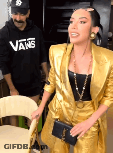 A Woman In A Gold Suit Is Standing Next To A Man Wearing A Vans Shirt GIF
