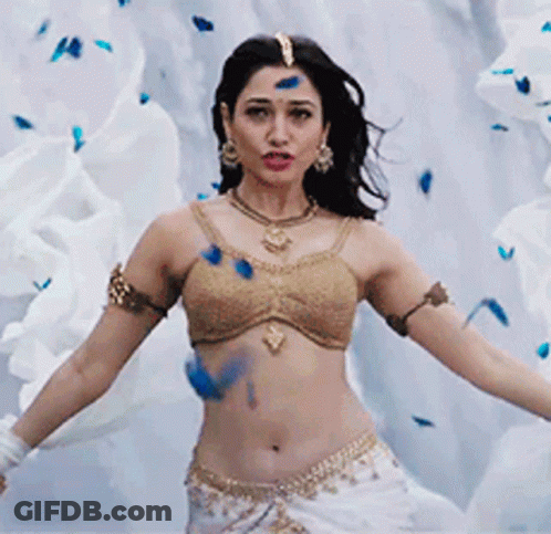 A Woman In A Gold Top And White Skirt Is Dancing In Front Of A White Wall . GIF
