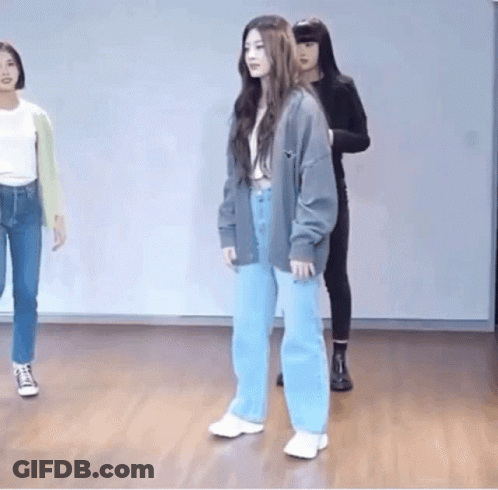 A Woman In A Gray Cardigan And Blue Jeans Is Standing On A Wooden Floor In A Dance Studio . GIF