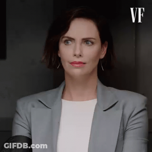 A Woman In A Gray Jacket And White Shirt Is Sitting In A Dark Room . GIF