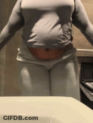 A Woman In A Gray Shirt And White Pants Is Standing In Front Of A Bathroom Sink . GIF