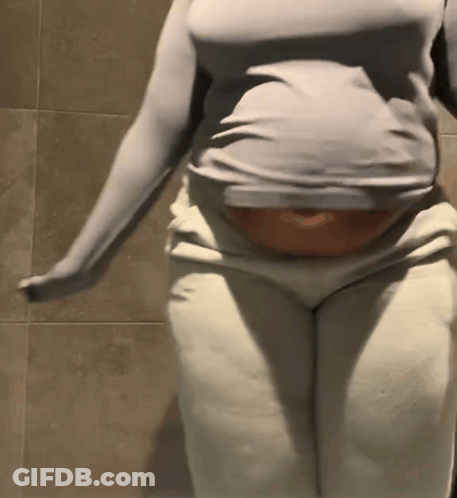 A Woman In A Gray Shirt And White Pants Is Standing In Front Of A Tiled Wall . GIF
