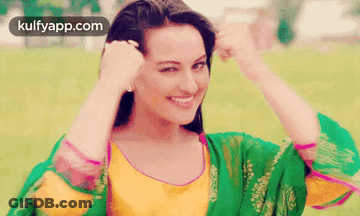 A Woman In A Green And Yellow Dress Is Smiling While Holding Her Hair . GIF