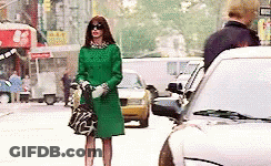 A Woman In A Green Coat And Sunglasses Is Standing On A Busy City Street . GIF