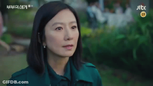 A Woman In A Green Coat Is Sitting In A Park Looking At The Camera . GIF