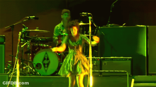 A Woman In A Green Dress Is Singing Into A Microphone In Front Of A Drum Set With The Letters Jbf On It GIF