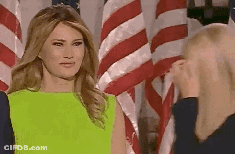 A Woman In A Green Dress Is Standing In Front Of An American Flag And Talking To Another Woman . GIF
