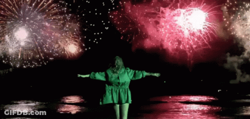 A Woman In A Green Dress Stands In Front Of A Firework Display GIF