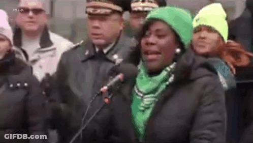 A Woman In A Green Hat Is Speaking Into A Microphone While Standing In Front Of A Crowd Of People . GIF