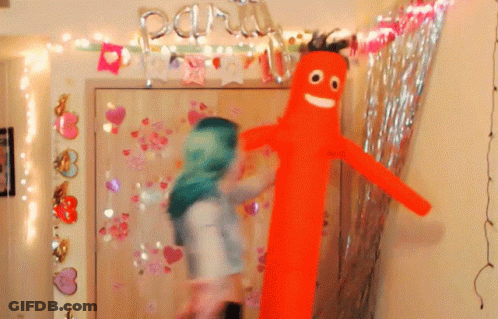 A Woman In A Green Head Scarf Stands Next To A Man In An Orange Balloon GIF
