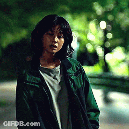 A Woman In A Green Jacket Is Standing On A Street . GIF