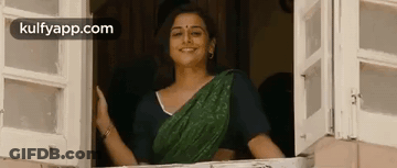 A Woman In A Green Sari Is Standing In A Window And Smiling . GIF