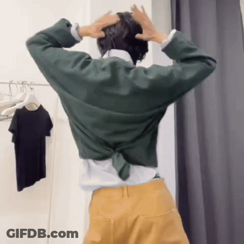 A Woman In A Green Shirt And Yellow Pants Is Standing In A Dressing Room With Her Hands On Her Head GIF