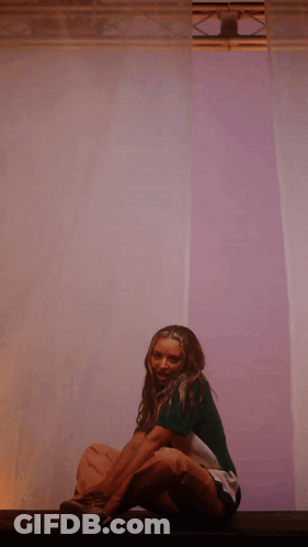 A Woman In A Green Shirt Sits On A Purple Wall GIF