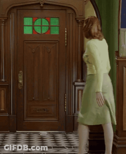 A Woman In A Green Skirt Is Walking In Front Of A Door With A Stained Glass Window GIF