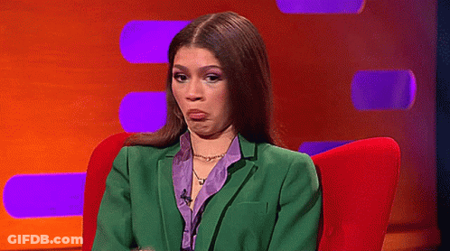 A Woman In A Green Suit And Purple Shirt Is Sitting In A Red Chair And Making A Funny Face . GIF