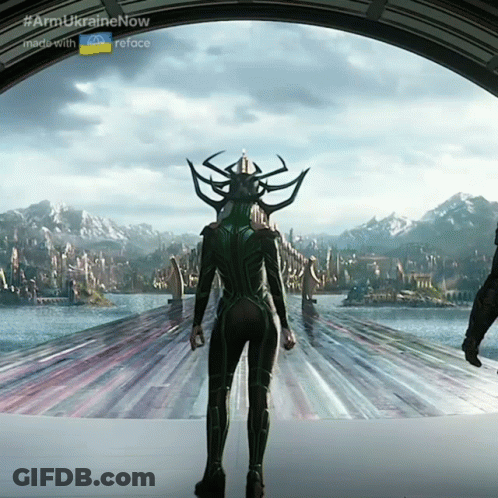 A Woman In A Green Suit Is Standing In Front Of A Body Of Water And Mountains GIF