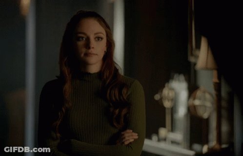 A Woman In A Green Sweater Stands With Her Arms Crossed In A Dark Room GIF