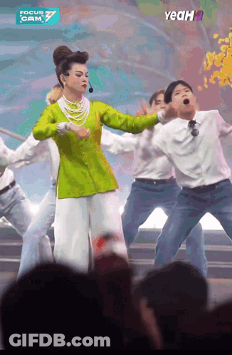 A Woman In A Green Top And White Pants Is Dancing In Front Of A Crowd And A Sign That Says Yeah GIF