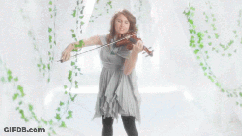 A Woman In A Grey Dress Is Playing A Violin In Front Of A White Curtain GIF