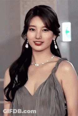 A Woman In A Grey Dress With A Necklace And Earrings Looks At The Camera GIF
