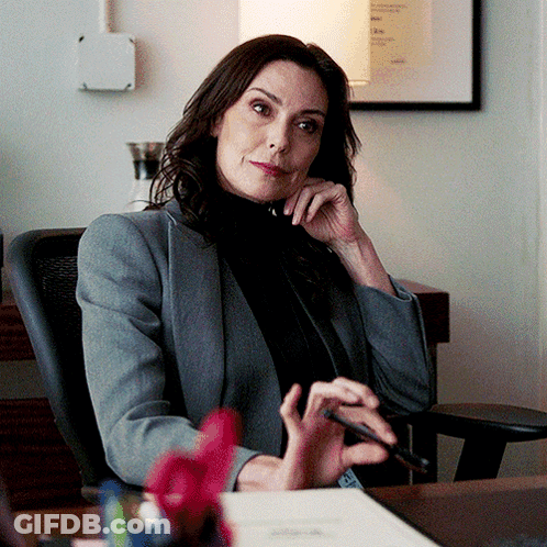 A Woman In A Grey Jacket Sits At A Desk With A Pen In Her Hand GIF