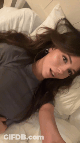 A Woman In A Grey Shirt Is Laying On A Bed With White Sheets GIF