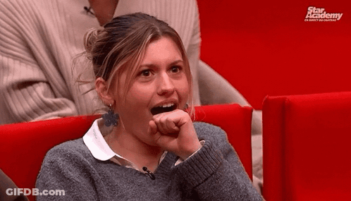 A Woman In A Grey Sweater Is Sitting In A Red Chair With A Star Academy Logo Behind Her GIF