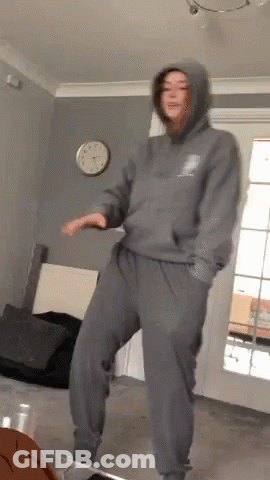A Woman In A Hoodie And Sweatpants Is Dancing In A Living Room GIF