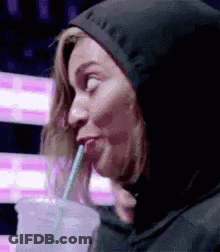 A Woman In A Hoodie Is Drinking Through A Straw From A Cup . GIF