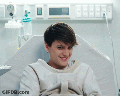 A Woman In A Hospital Gown Is Smiling And Looking At The Camera While Sitting In A Bed GIF