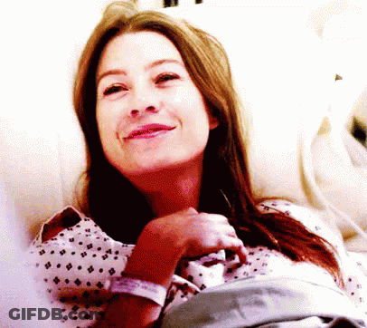 A Woman In A Hospital Gown Smiles While Laying In A Hospital Bed GIF