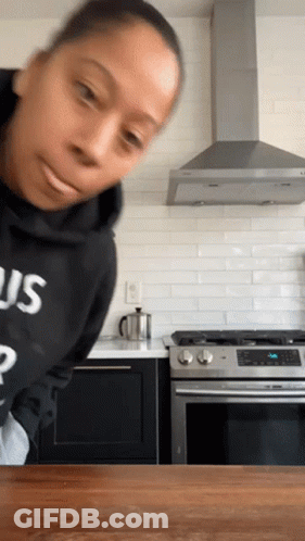 A Woman In A Kitchen Wearing A Black Sweatshirt With The Word Us On It GIF