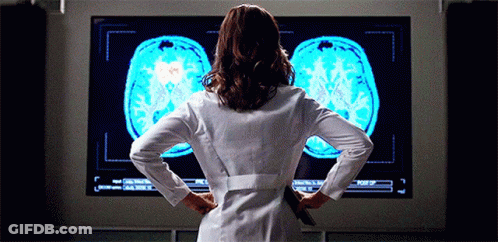 A Woman In A Lab Coat Stands In Front Of A Large Screen Showing A Brain Scan GIF