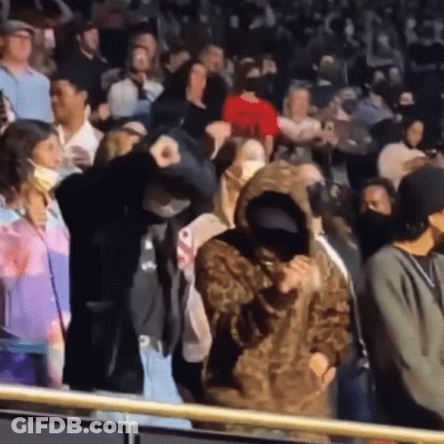 A Woman In A Leopard Print Hoodie Stands In A Crowd Of People Wearing Face Masks GIF