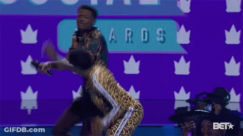 A Woman In A Leopard Print Outfit Stands Next To A Man Holding A Microphone At An Awards Show GIF