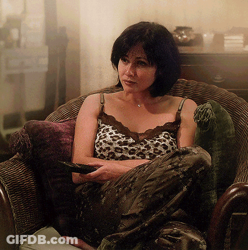 A Woman In A Leopard Print Tank Top Sits In A Chair Holding A Remote Control GIF