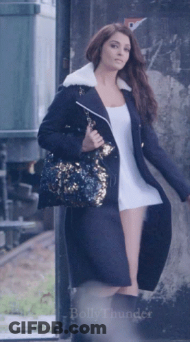 A Woman In A Long Black Coat Is Holding A Purse And The Word Bollythunder Is On The Bottom Of The Image GIF