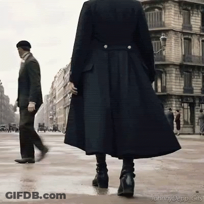 A Woman In A Long Black Coat Is Walking Down A Street While A Man In A Suit Walks Behind Her . GIF