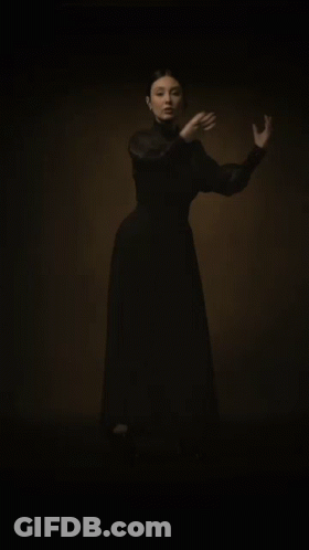 A Woman In A Long Black Dress Is Standing In The Dark With Her Hands Outstretched . GIF