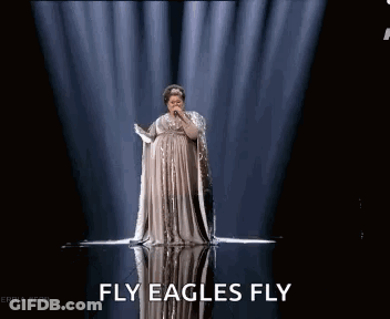 A Woman In A Long Dress Is Singing Into A Microphone On A Stage With The Words Fly Eagles Fly Above Her GIF