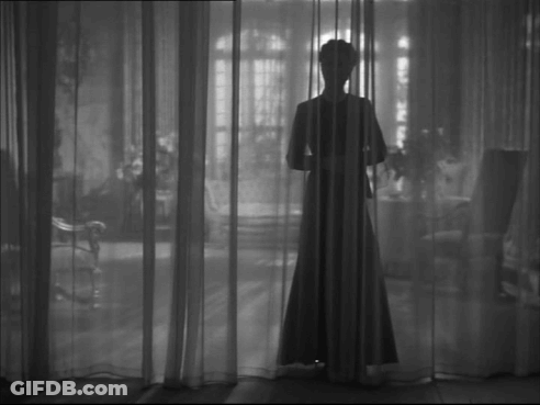 A Woman In A Long Dress Is Standing Behind A Curtain In A Black And White Photo GIF
