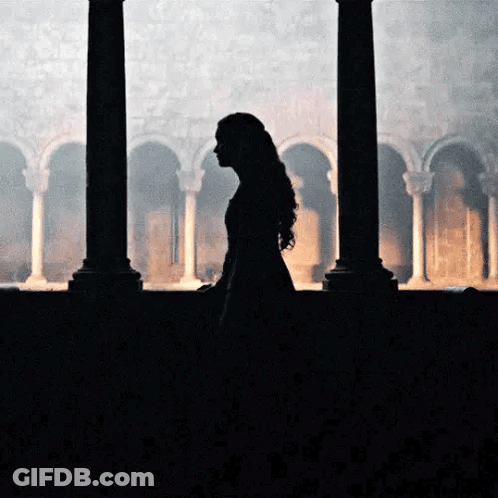 A Woman In A Long Dress Is Standing In Front Of A Window In A Dark Room . GIF