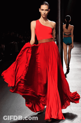 A Woman In A Long Red Dress Is Walking Down A Runway GIF