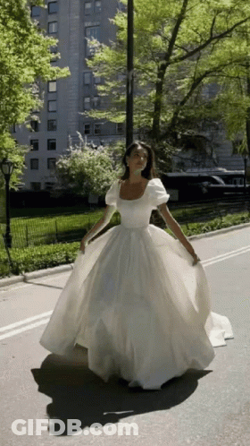A Woman In A Long White Dress Is Walking Down A Street GIF